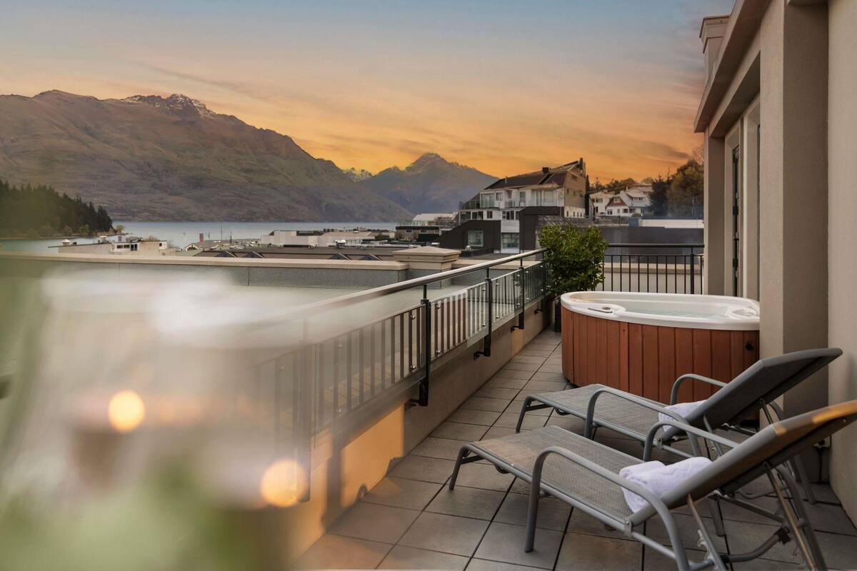 A balcony with a hot tub, lounge chairs, and a view of a lake and mountains.