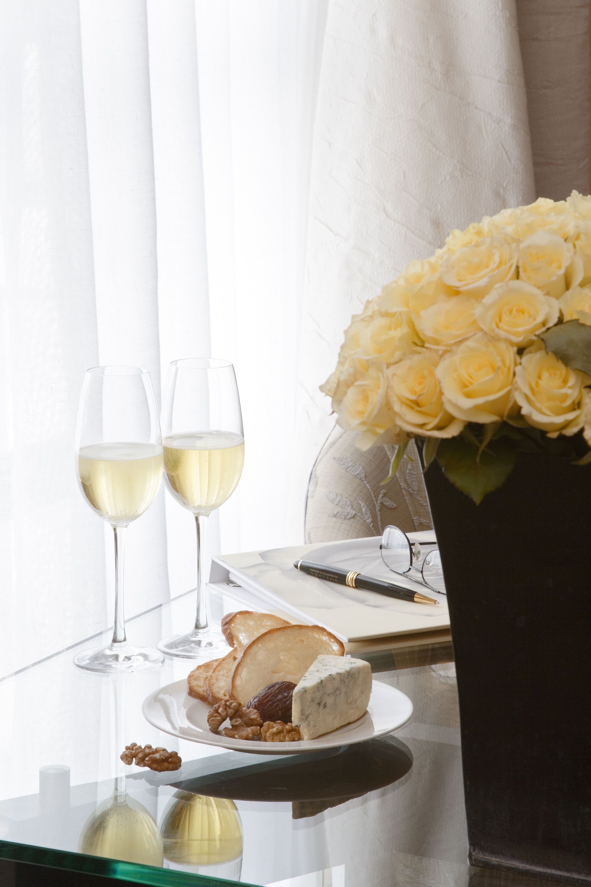 A table with two glasses of white wine, a plate with cheese and nuts, a vase of yellow roses, and a pen on a notepad.