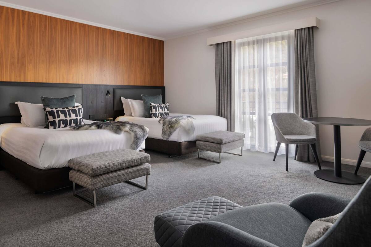A hotel room with two beds, a small table, a chair, and a view of the city through large windows.