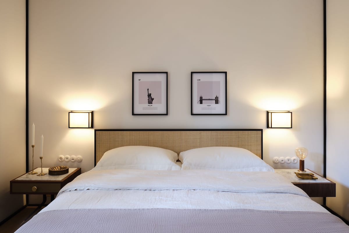 A neatly made bed with a headboard, two bedside tables with wall-mounted lamps, and two framed pictures on the wall.