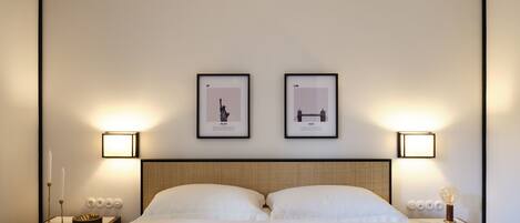 Un lit bien fait, avec une tête de lit, deux tables de chevet dotées de lampes fixées au mur, et deux tableaux encadrés accrochés au mur.