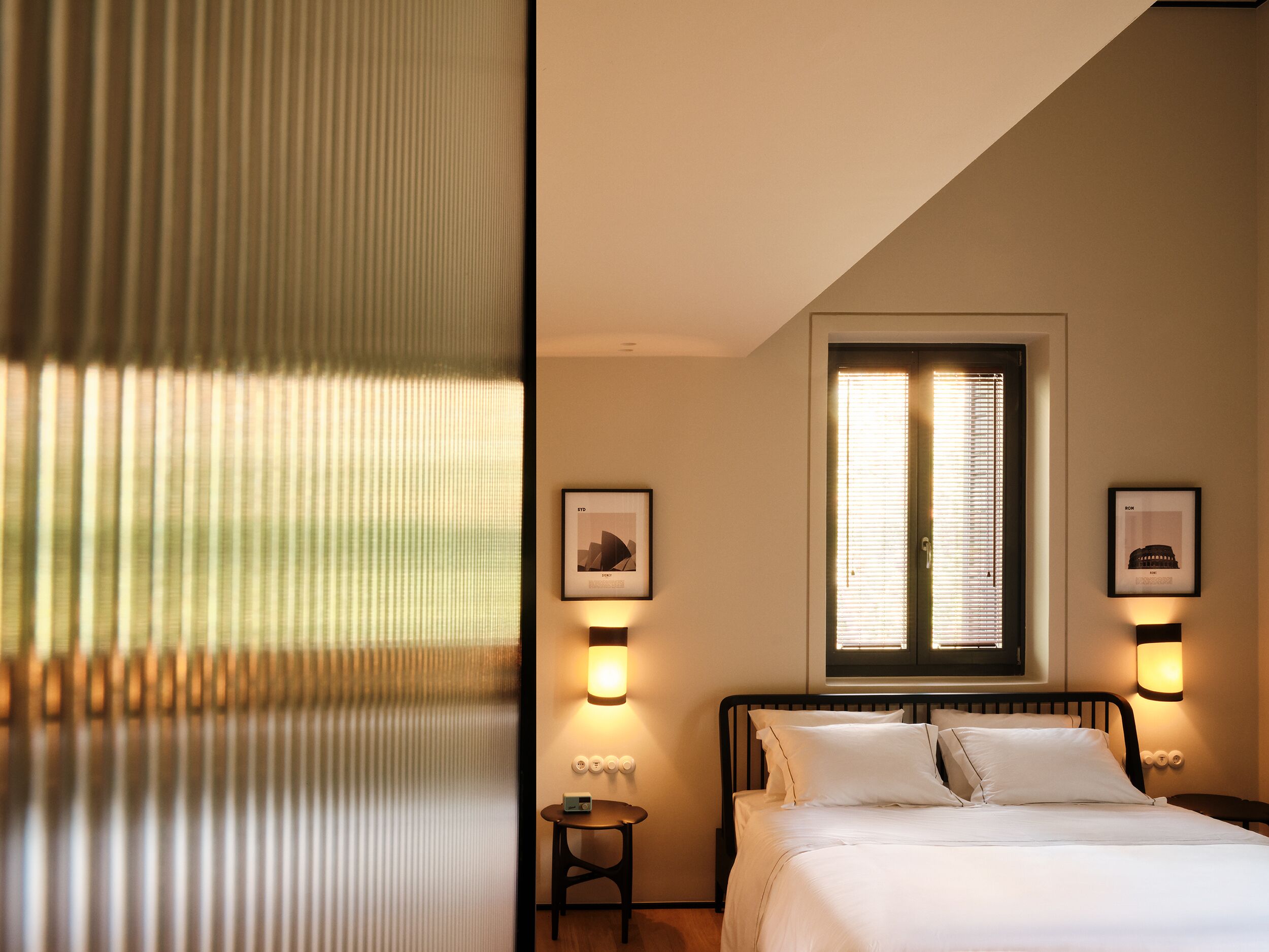 A bedroom with a bed, a window with blinds, two wall-mounted light fixtures, and a framed picture on the wall.