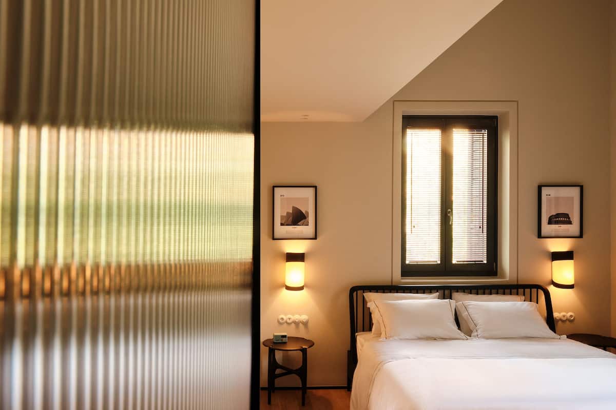A bedroom with a bed, a window with blinds, two wall-mounted light fixtures, and a framed picture on the wall.