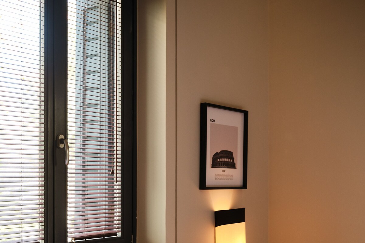 A hotel room with a bed, a nightstand, a framed picture, and a window with blinds.