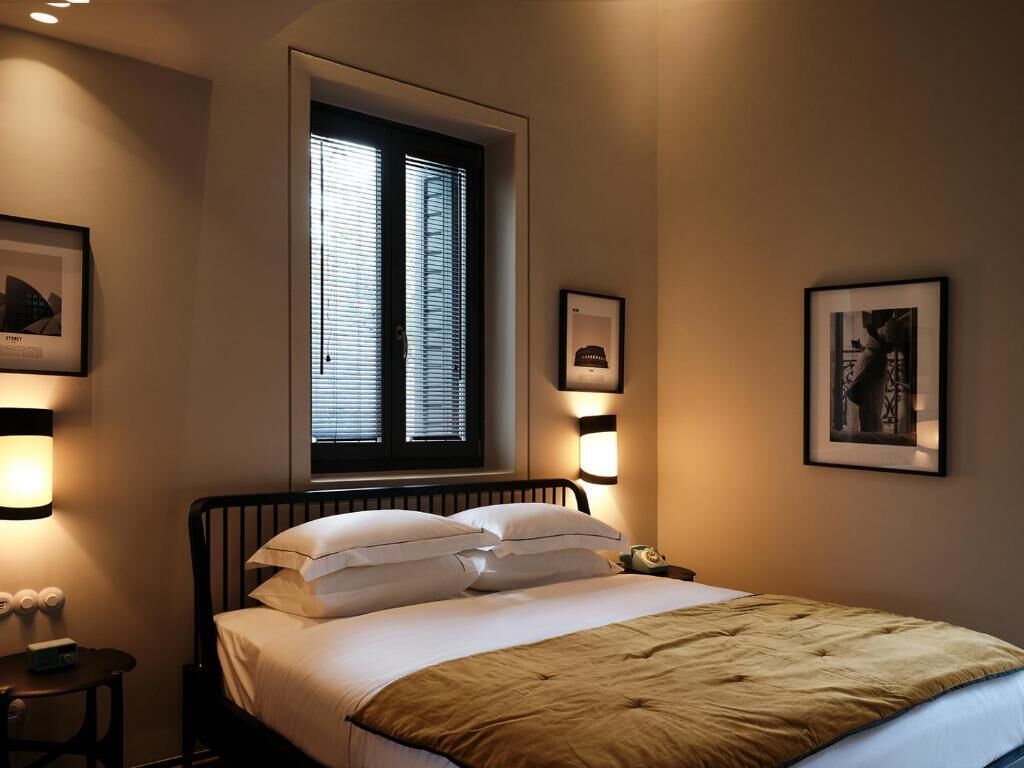 A bedroom with a bed, a nightstand, a window with blinds, and wall-mounted lights.