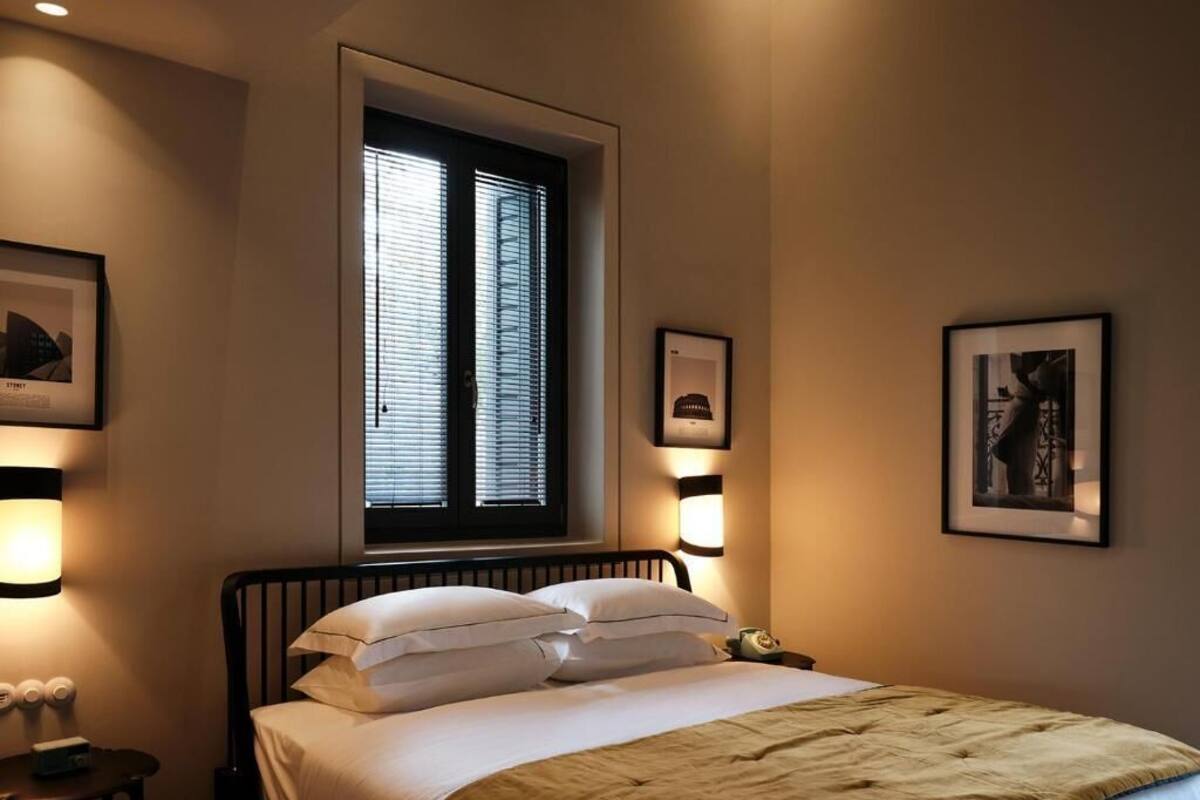 Une chambre à coucher comprenant un lit, une table de chevet, une fenêtre avec des stores et des lampes fixées au mur.
