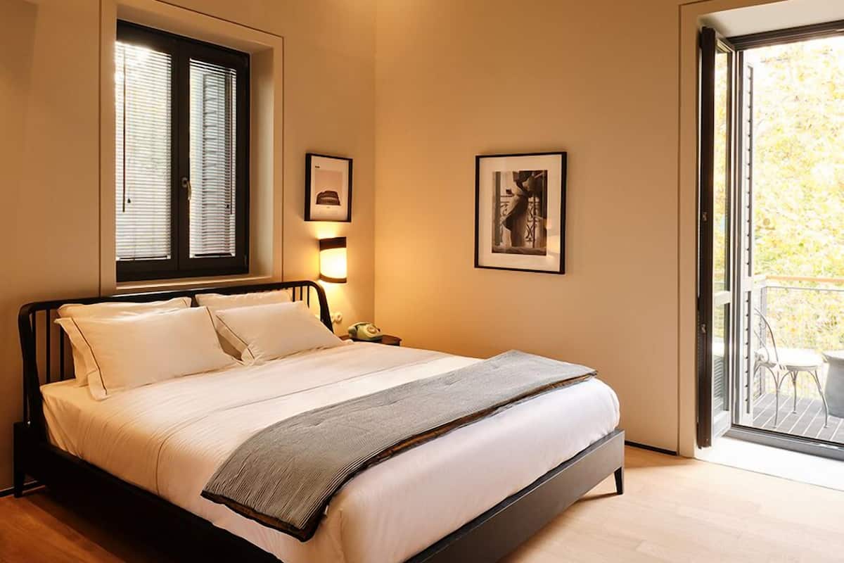 A modern bedroom with a bed, a nightstand, a window with blinds, and a door leading to a balcony.