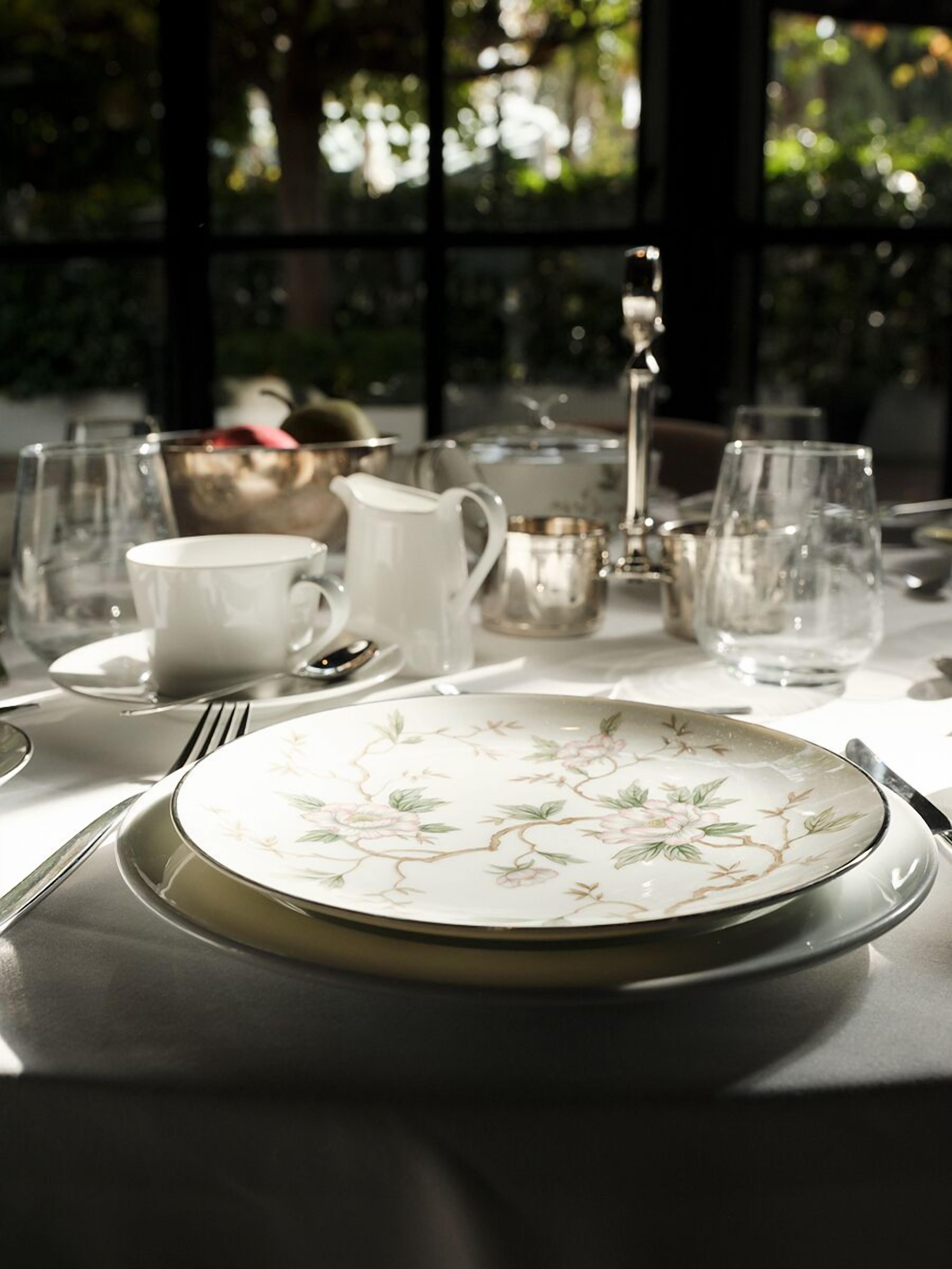 A table set with a floral plate, a white pitcher, and various glassware, with a view of greenery through a window.