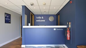 Ein Empfangsschalter mit einer Uhr und einem Schild mit der Aufschrift 'hello & welcome', ein Feuerlöscher und das 'Travelodge'-Logo.