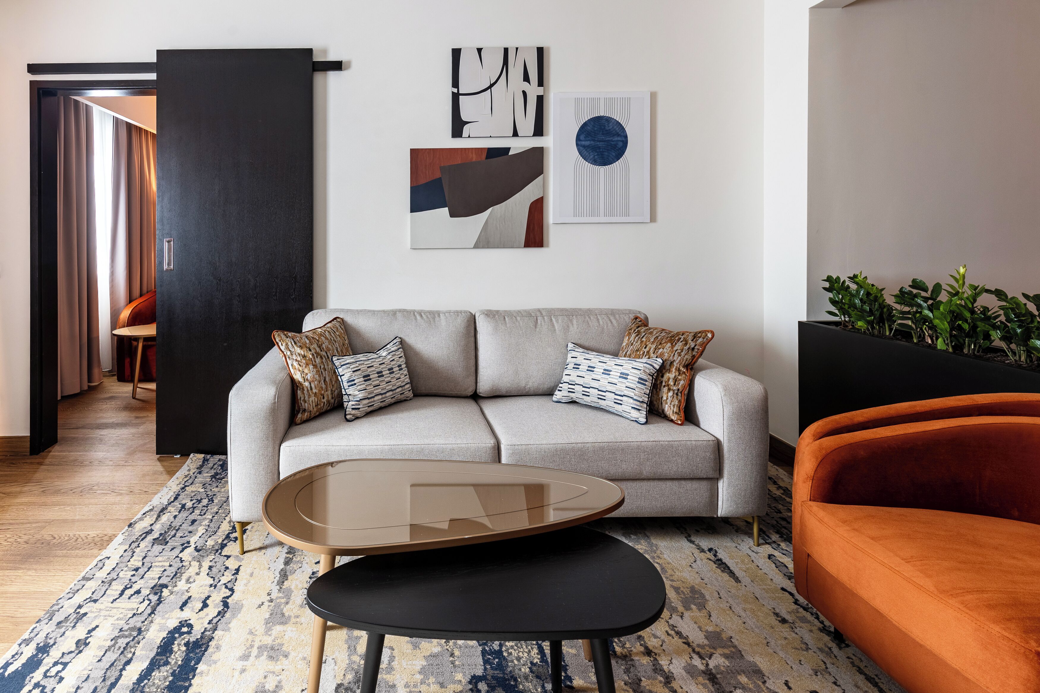 A modern living room with a grey sofa, a round glass coffee table, and an orange armchair. There are abstract wall art pieces above the sofa.