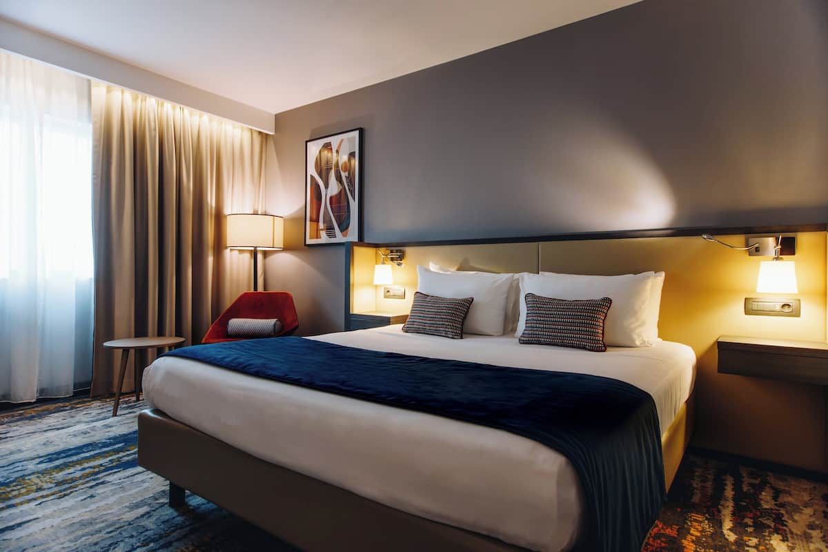 Une chambre d’hôtel avec un grand lit, deux lampes de chevet, une tête de lit, un fauteuil rouge, une petite table et une grande fenêtre avec des rideaux.
