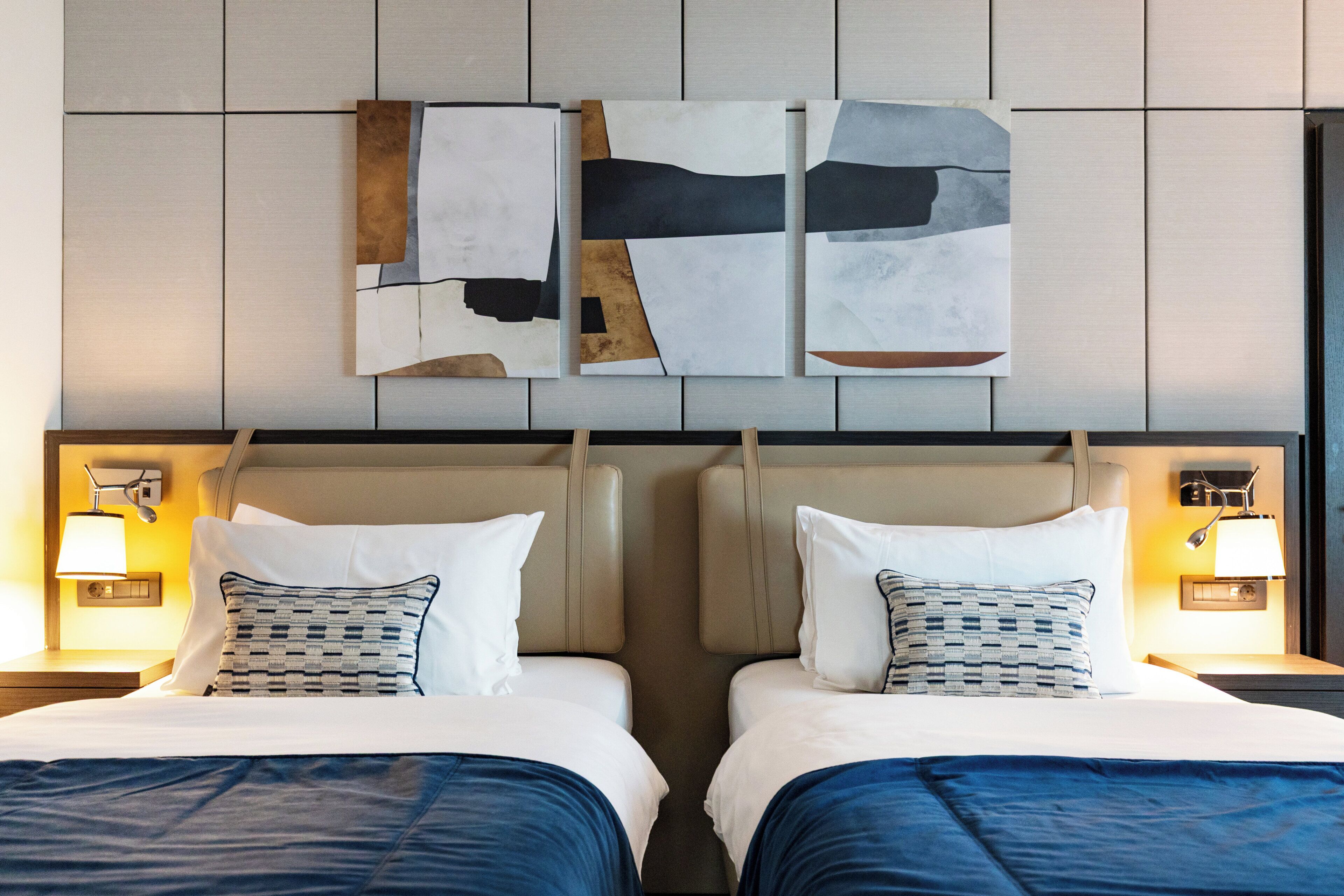 A hotel room with two beds, each with a headboard, a nightstand with a lamp, and abstract wall art above the beds.