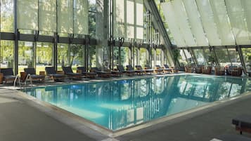 An indoor swimming pool with clear glass walls, sun loungers, and a view of trees outside.