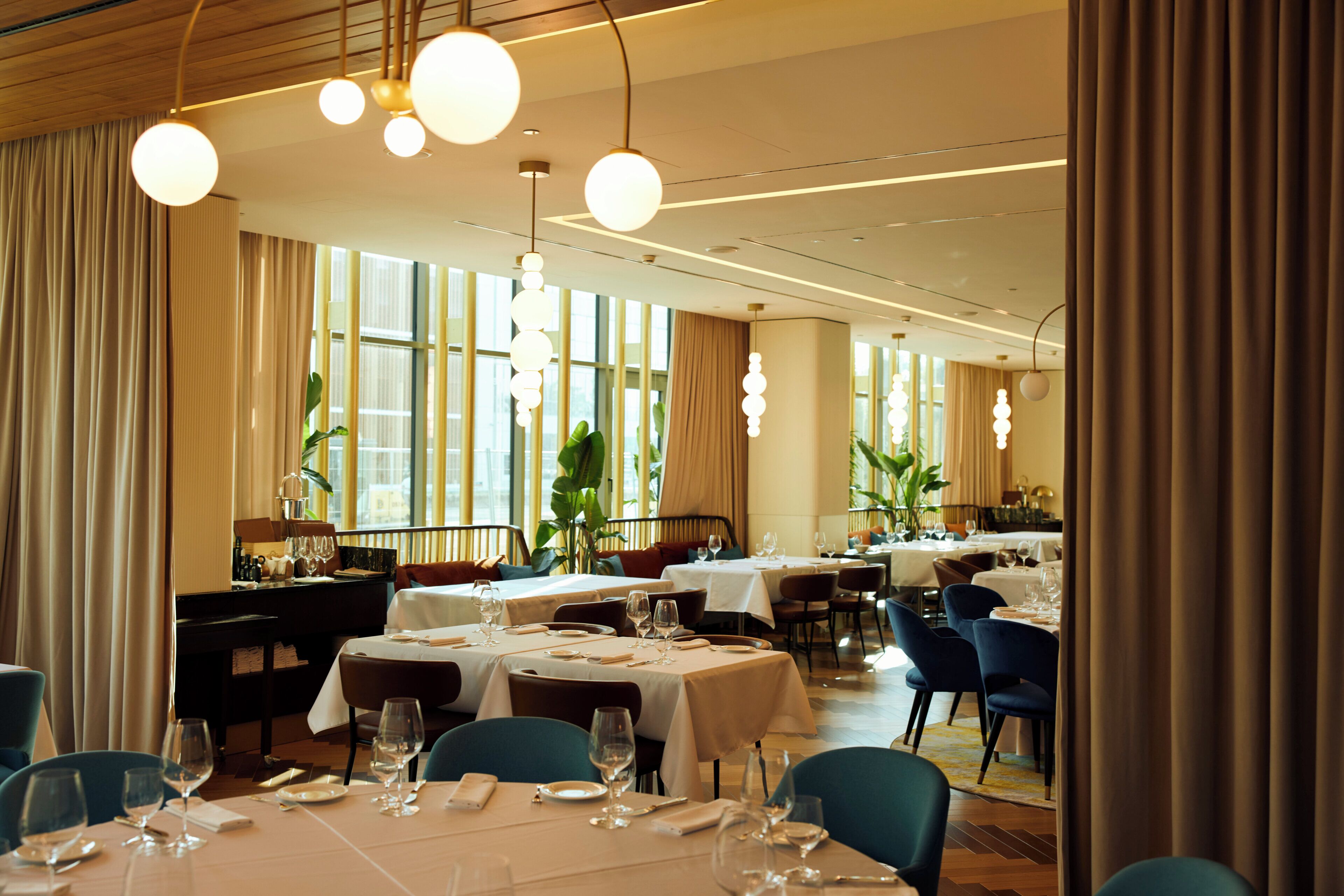 A spacious dining area with multiple tables set for dining, large windows allowing natural light, and hanging spherical light fixtures.