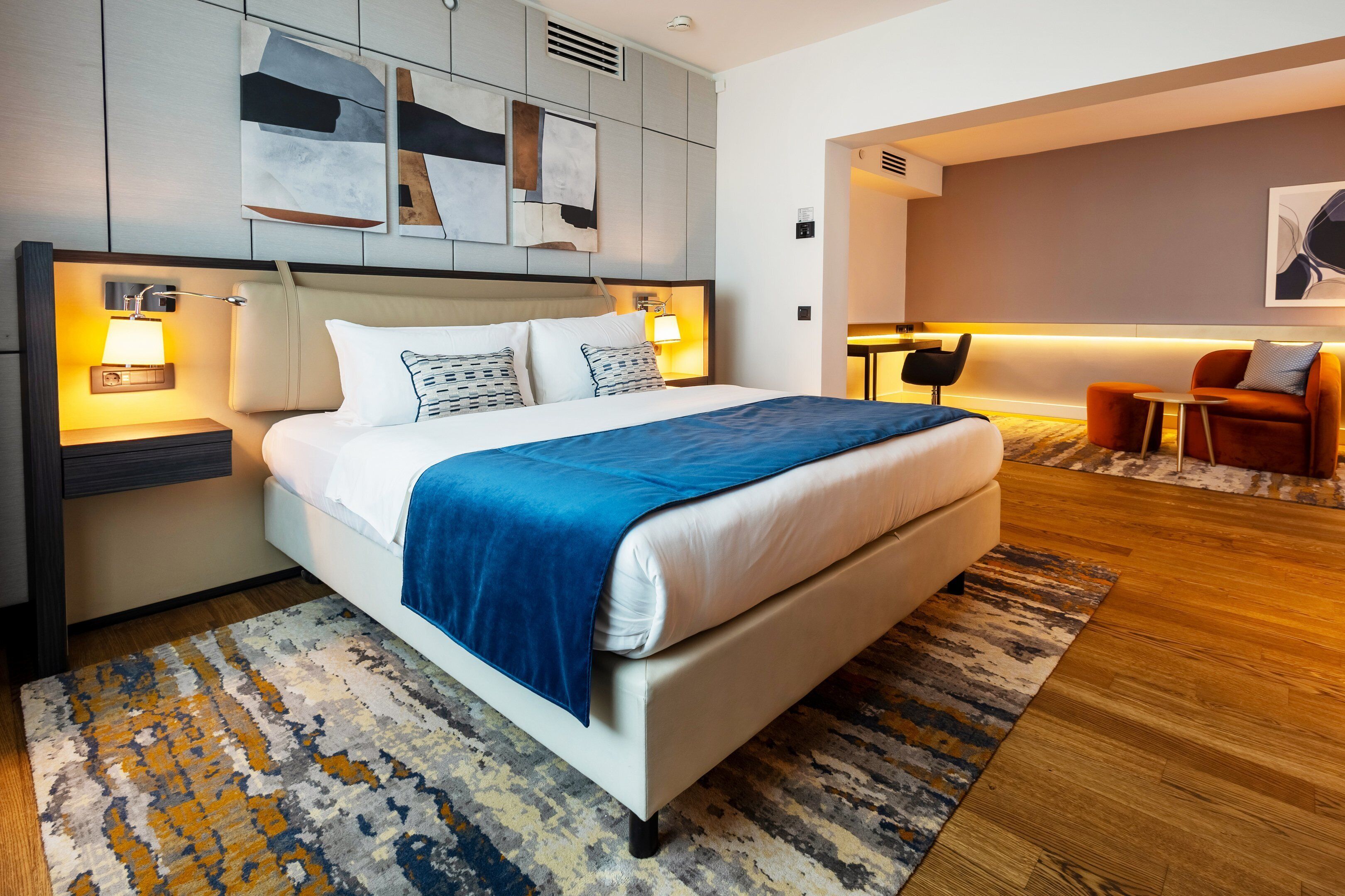 A modern hotel room with a large bed, two bedside tables with lamps, a headboard with abstract art, a seating area with a sofa and chairs, and a patterned rug.