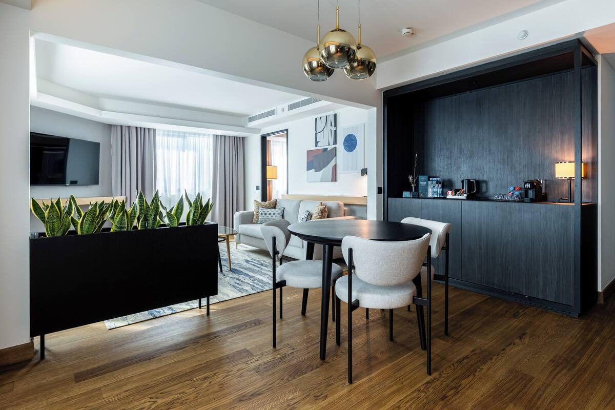 Ein modernes Hotelzimmer mit Essbereich, einem flachbildfernseher an der Wand, einer Couch, einer Kaffeestation und einem großen Fenster mit Vorhängen.