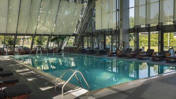 An indoor swimming pool with clear glass walls, sun loungers, and a tiled floor.