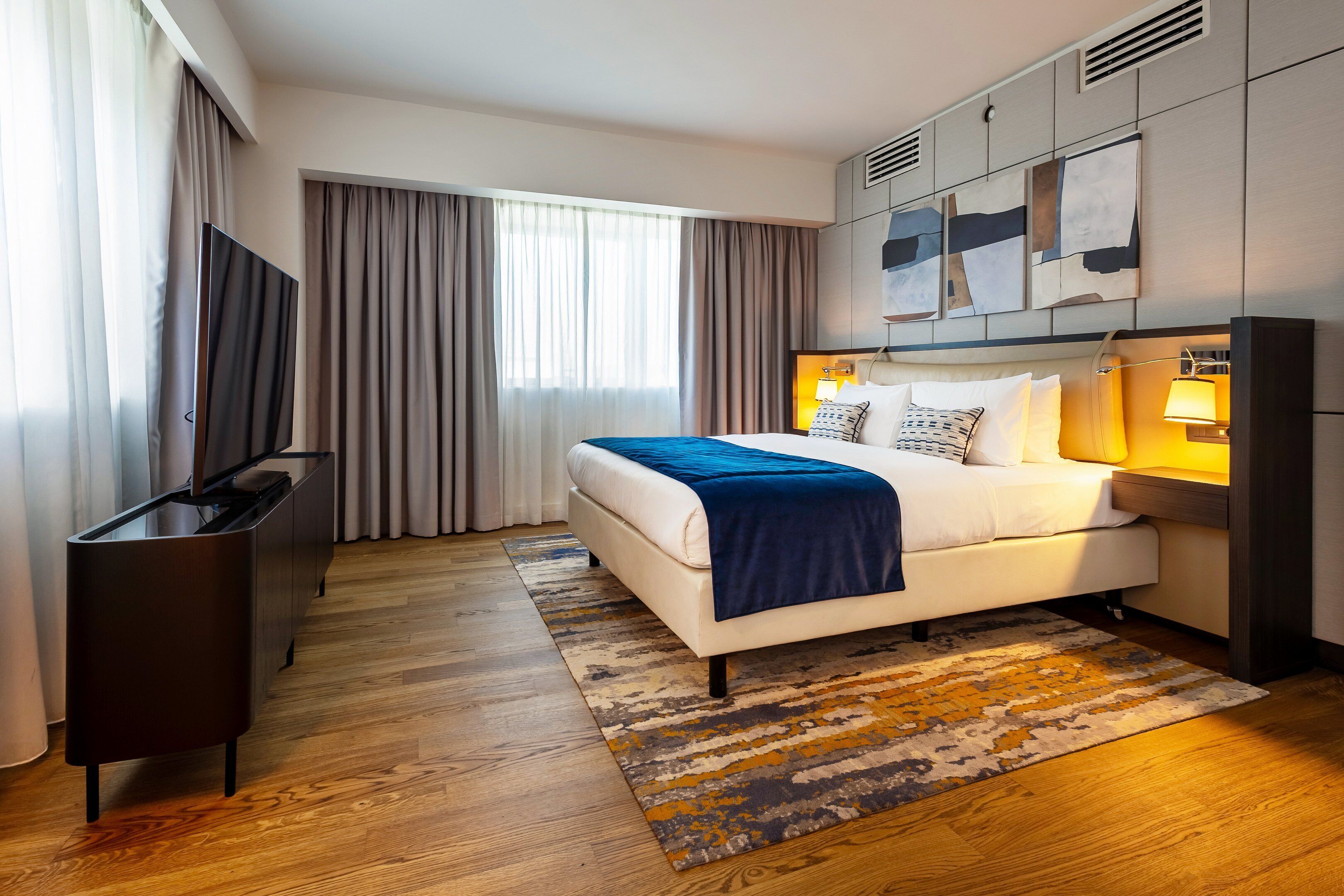 Una habitación de hotel moderna con una cama grande, un televisor de pantalla plana, un escritorio y un ventanal con cortinas.