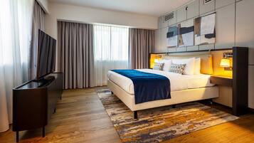Una habitación de hotel moderna con una cama grande, un televisor de pantalla plana, un escritorio y un ventanal con cortinas.