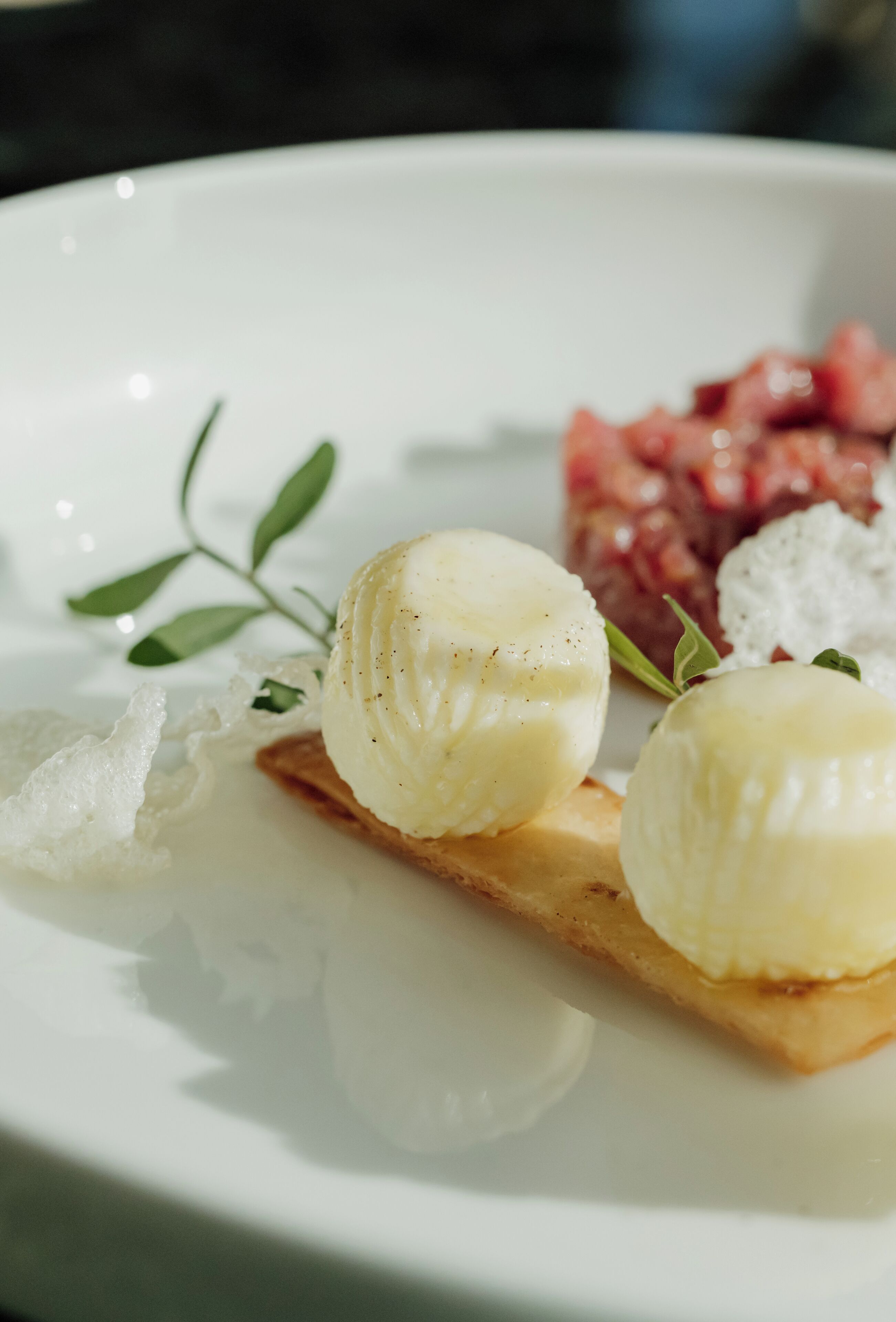 Ein Teller mit zwei runden, weißen, cremigen Desserts auf einer rechteckigen Unterlage, begleitet von einer kleinen Portion rosa Nahrung und einer weißen, krümeligen Masse.