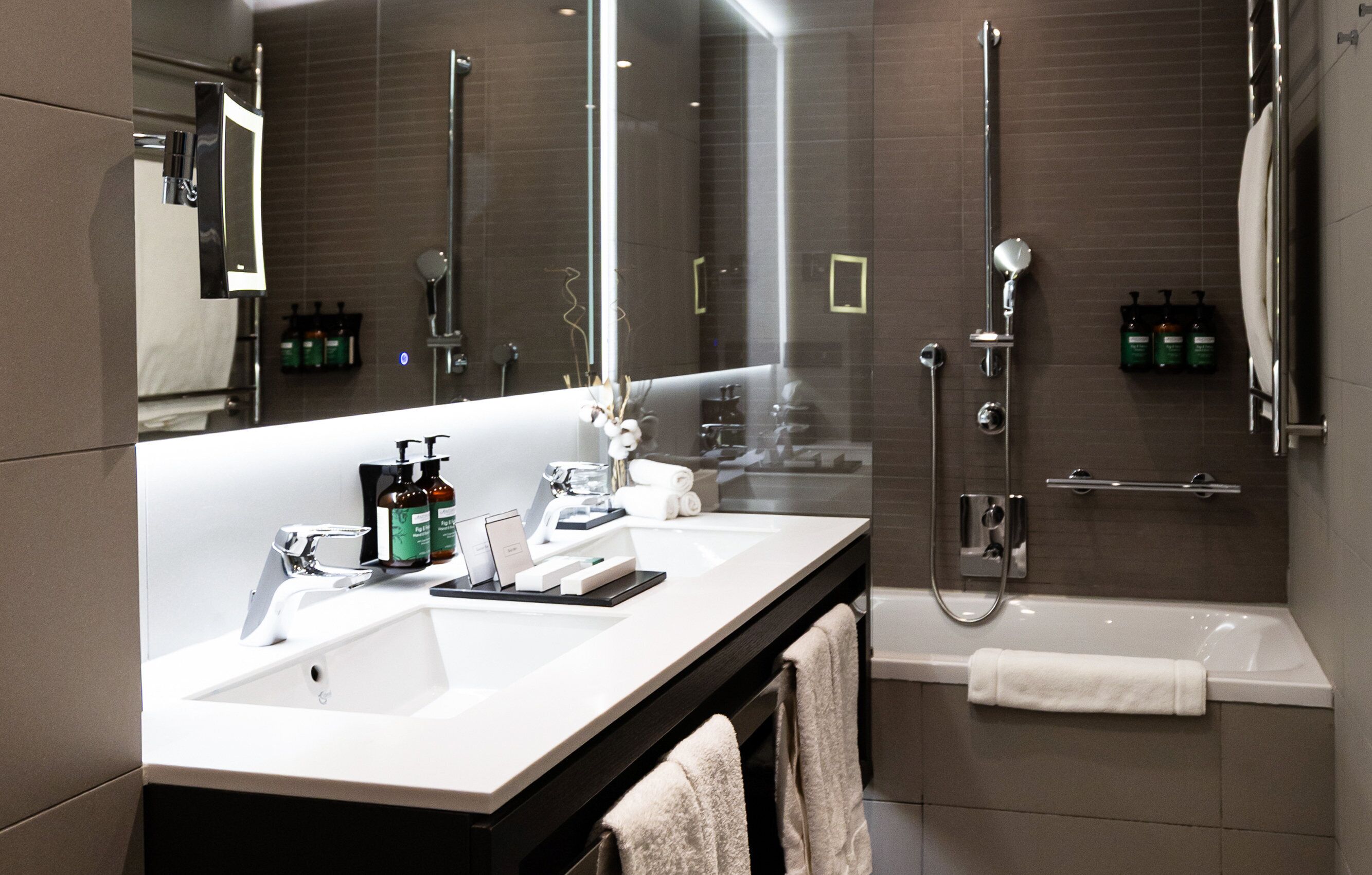 Ein modernes Badezimmer mit einem großen Spiegel, einem weißen Waschbecken, einer Dusche und einer Badewanne. Auf dem Waschbecken stehen Pflegeprodukte und ein Handtuch hängt an der Wand.