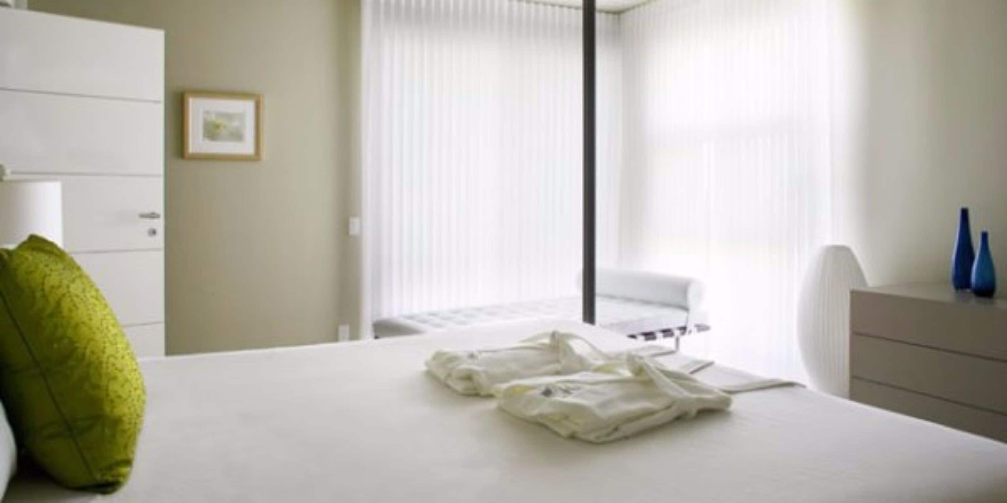Una camera d'albergo con un letto, un cuscino verde, un comodino bianco con una lampada e una finestra con persiane.