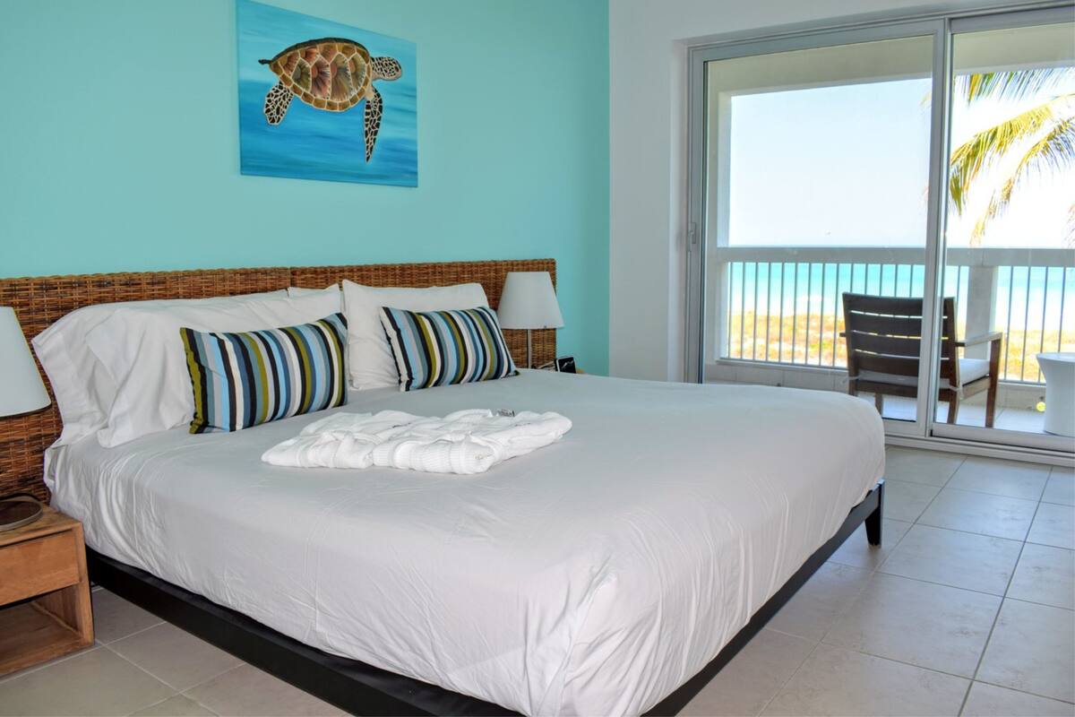 Ein Schlafzimmer mit einem großen Bett, zwei Kissen, einer weißen Decke, einem Nachttisch, einer Lampe und Blick auf den Strand durch eine Schiebetür.