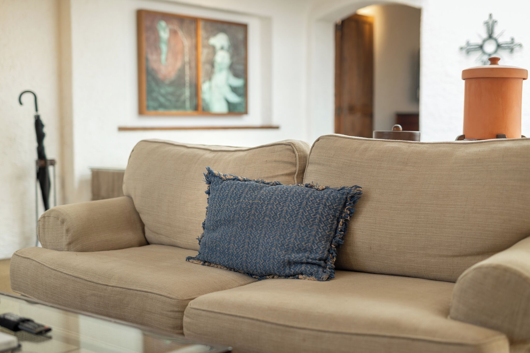 Un sofá color beige con un cojín azul con textura y una mesa de centro de vidrio con un control remoto.