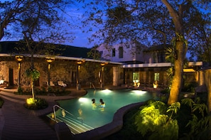 A pool area with two people swimming, surrounded by a stone building with outdoor seating and a view of the sky.