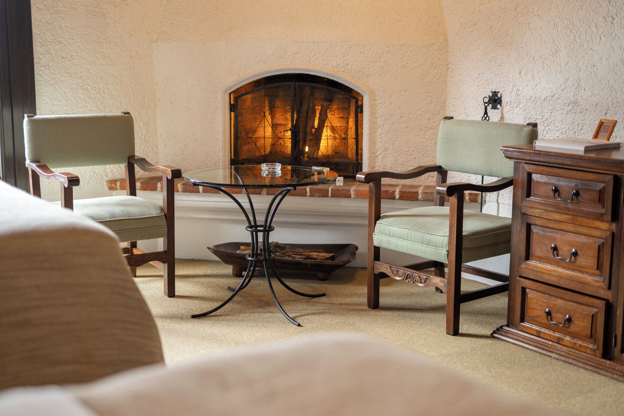 A cozy room with a fireplace, two armchairs, a wooden dresser, and a glass table.