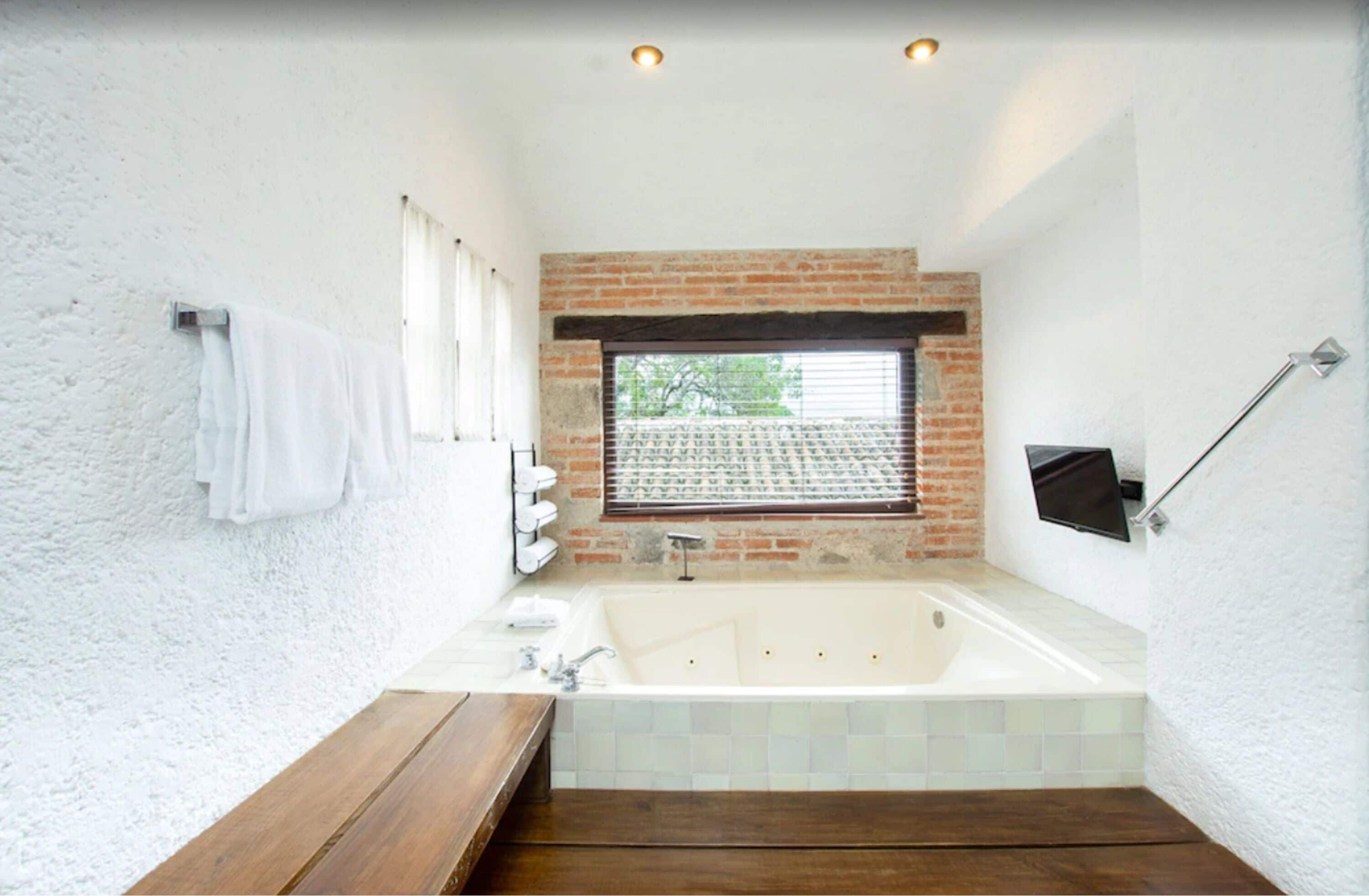 A modern bathroom with a large bathtub, a wooden bench, a towel rack, and a window with blinds.