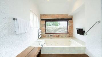 A modern bathroom with a large bathtub, a wooden bench, a towel rack, and a window with blinds.