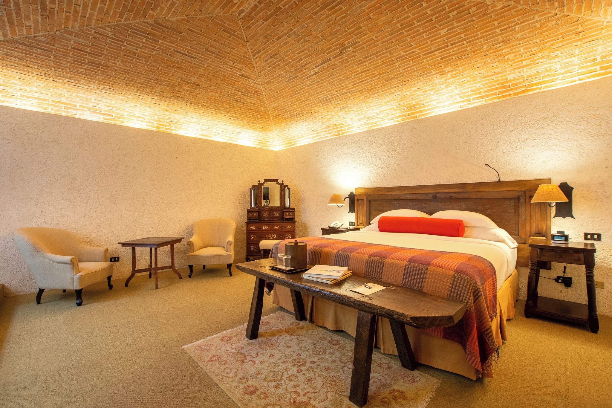 A cozy hotel room with a brick ceiling, a large bed with a red pillow, two armchairs, a wooden nightstand, and a small table with a lamp.