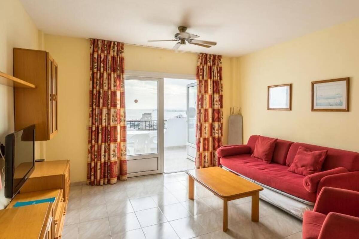 Un soggiorno con un divano rosso, un tavolino in legno, una televisione e una ventola a soffitto. Si vedono tende, una porta che dà su un balcone e due quadri appesi al muro.