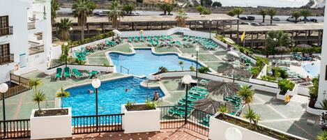 A rooftop pool area with multiple swimming pools, lounge chairs, and palm trees.