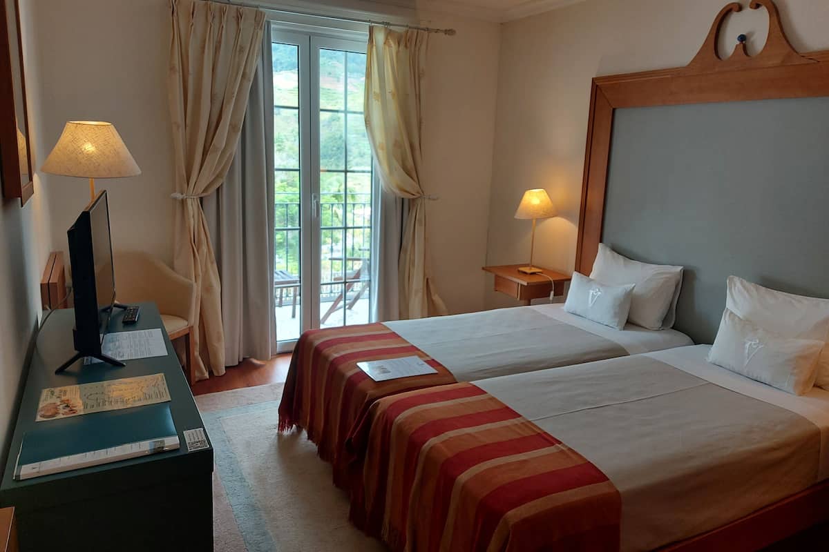 Een hotelkamer met twee bedden, een bureau met een lamp, een raam met gordijnen en uitzicht op groen buiten.