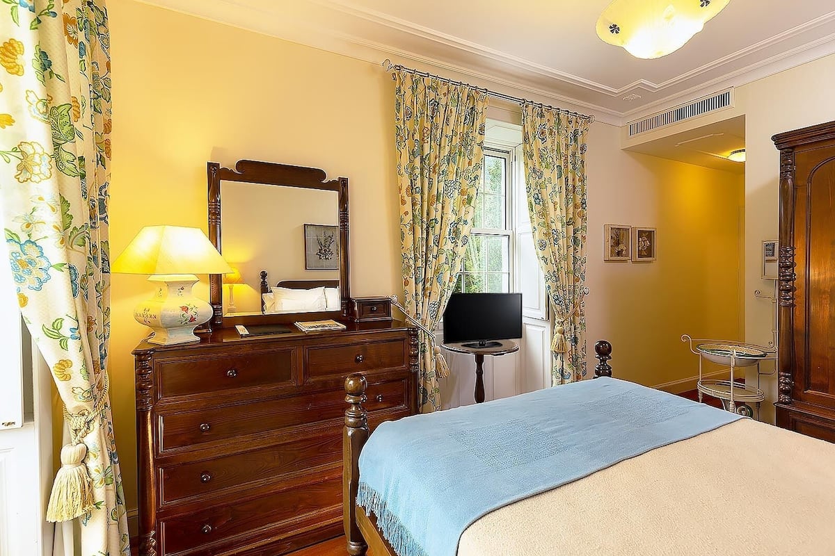 Een slaapkamer met een houten dressoir, een bed met een blauwe deken, een nachtkastje met een lamp, een televisie en bloemen gordijnen.
