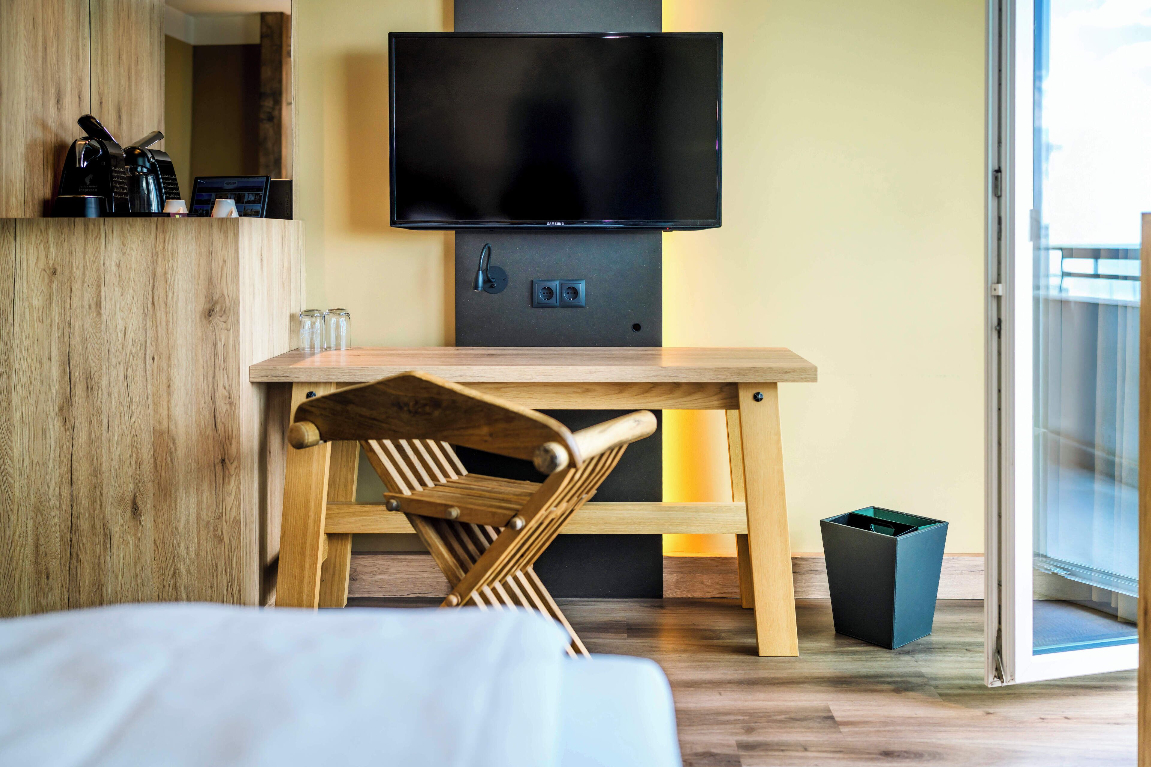 Une chambre d’hôtel moderne avec un bureau en bois, un téléviseur fixé au-dessus, et une porte-fenêtre coulissante donnant sur un balcon.
