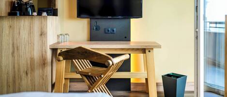 Una habitación de hotel moderna con un escritorio de madera, un televisor montado encima y una puerta corredera de vidrio que da a un balcón.