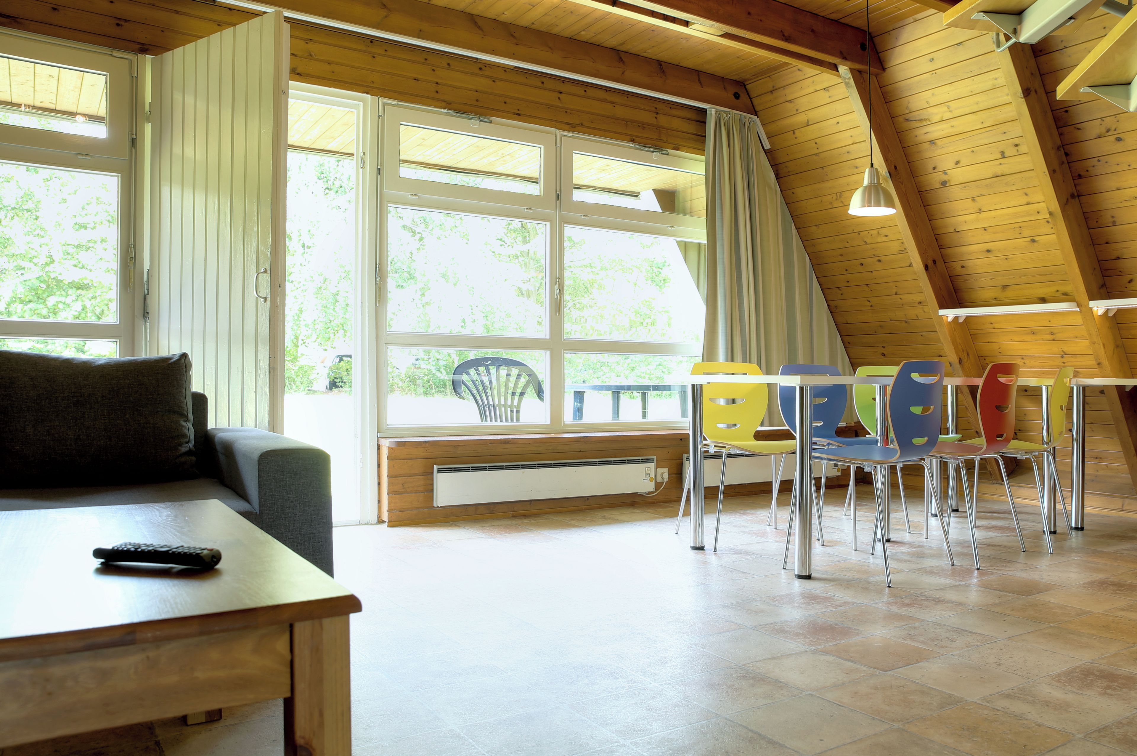 A room with a wooden ceiling, a sofa, a dining table with chairs, and large windows.