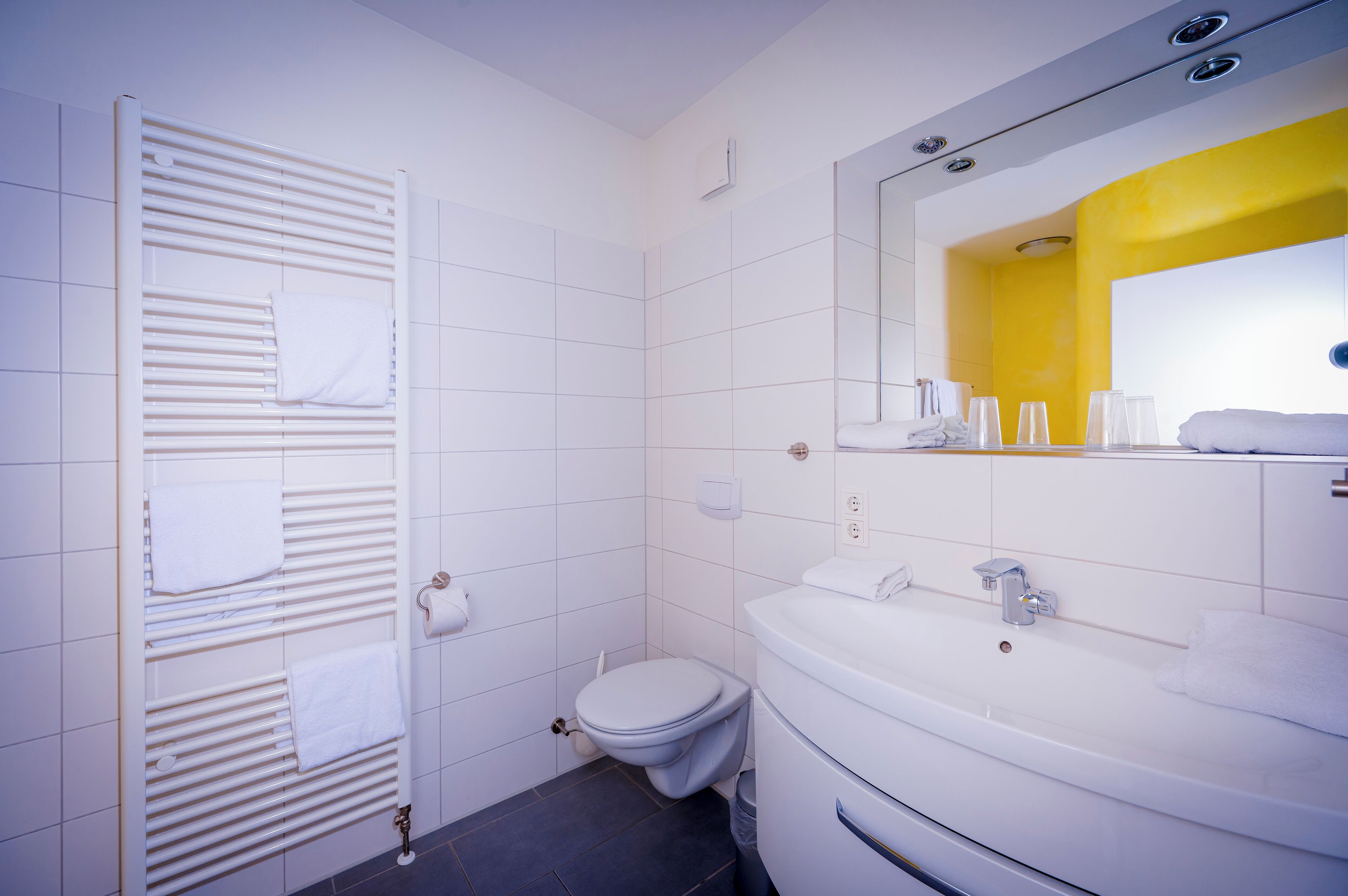 Ein modernes Badezimmer mit einer weißen Toilette, einem Waschbecken inklusive Spiegel und einem Handtuchhalter mit mehreren Handtüchern.