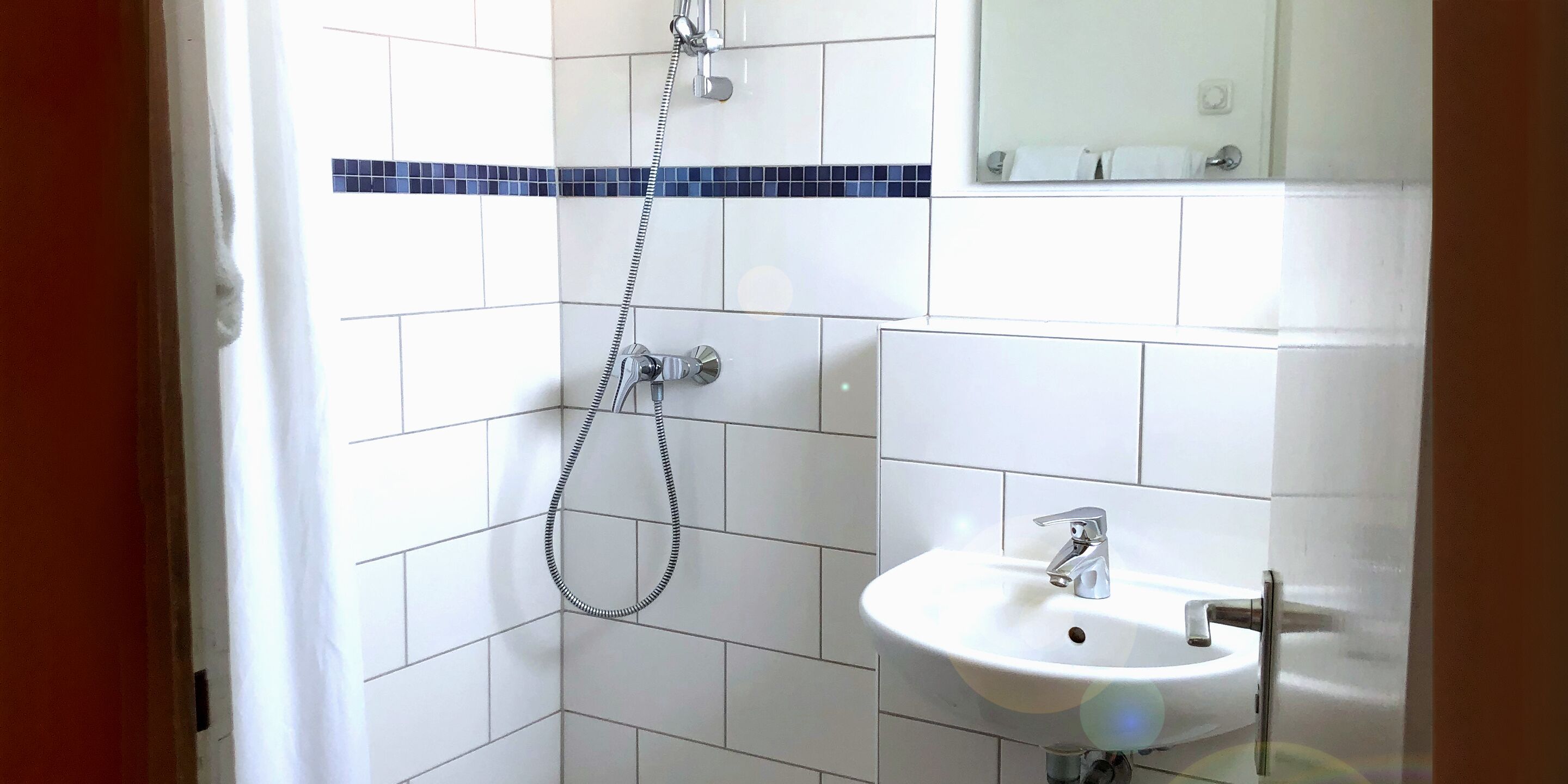 A bathroom with white tiled walls, a shower with a handheld hose, and a sink with a chrome faucet.