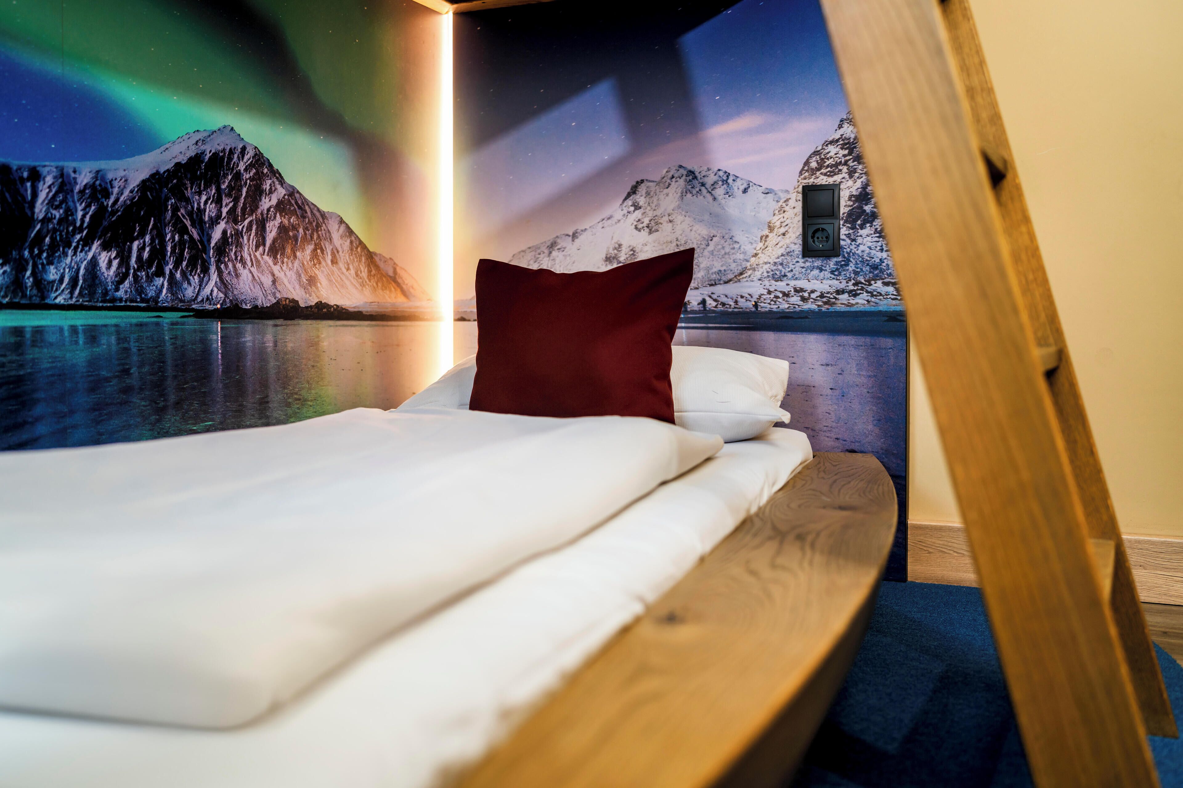 A bed with white bedding and a red pillow, set against a wall mural depicting a snowy mountain and a lake under a starry sky.