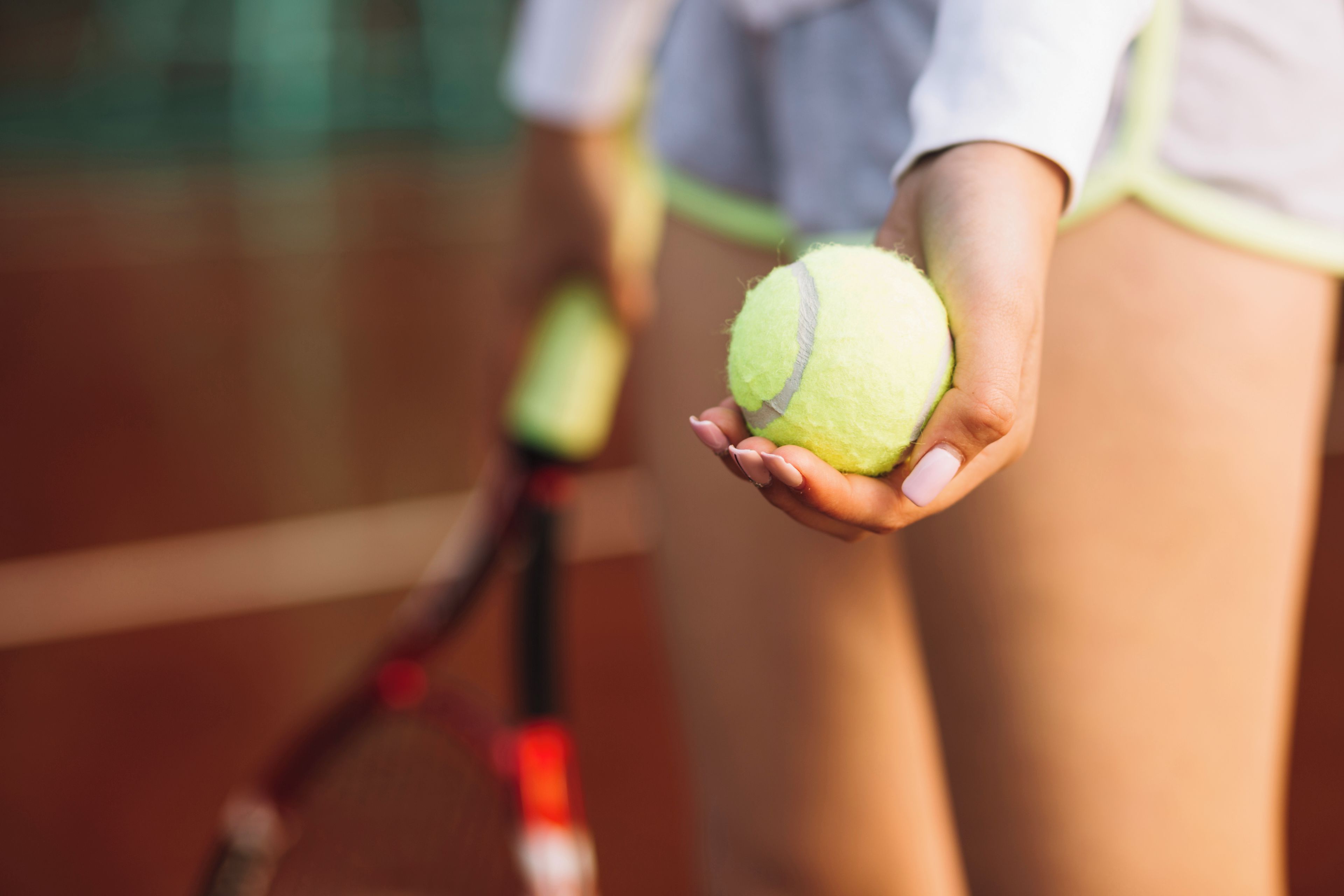 Nahaufnahme eines Tennisballs in einer Hand, ein Tennisracket ist im Hintergrund zu sehen.