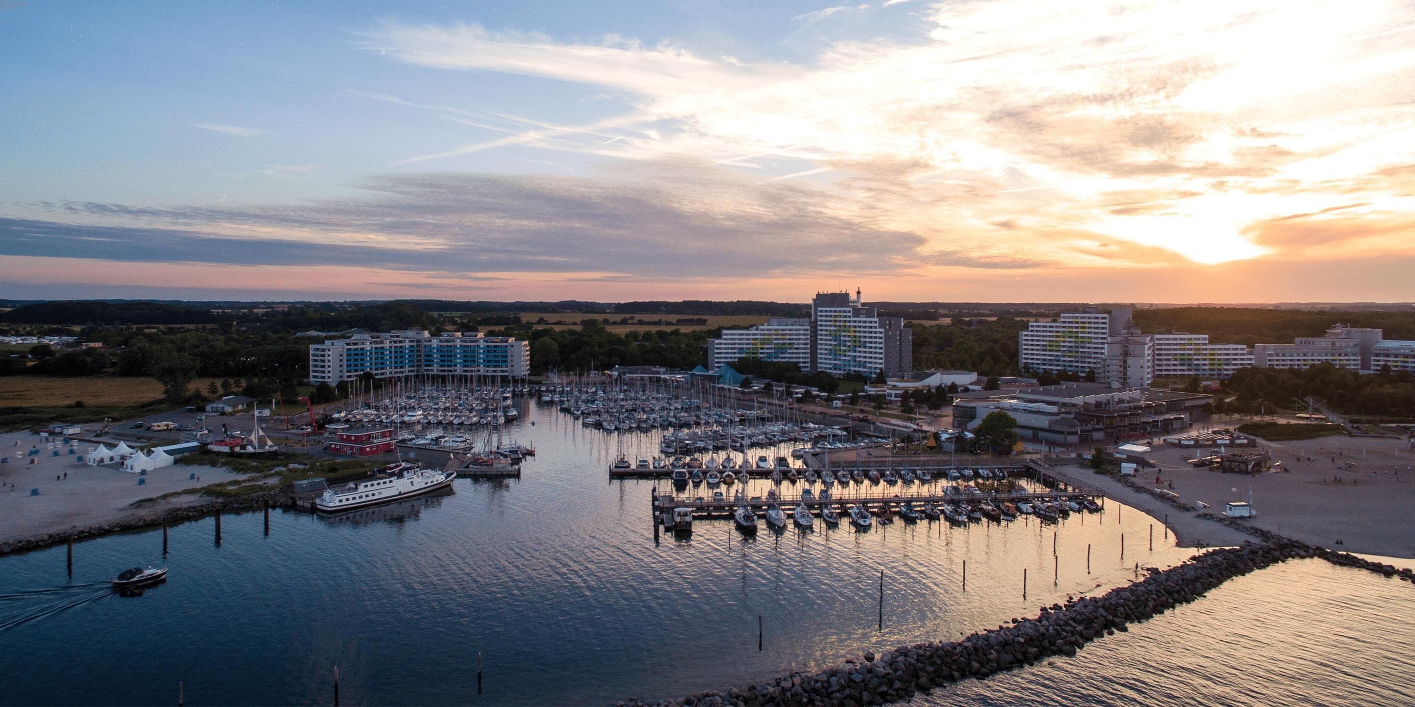Un puerto deportivo con barcos amarrados, rodeado de edificios y una puesta de sol al fondo.