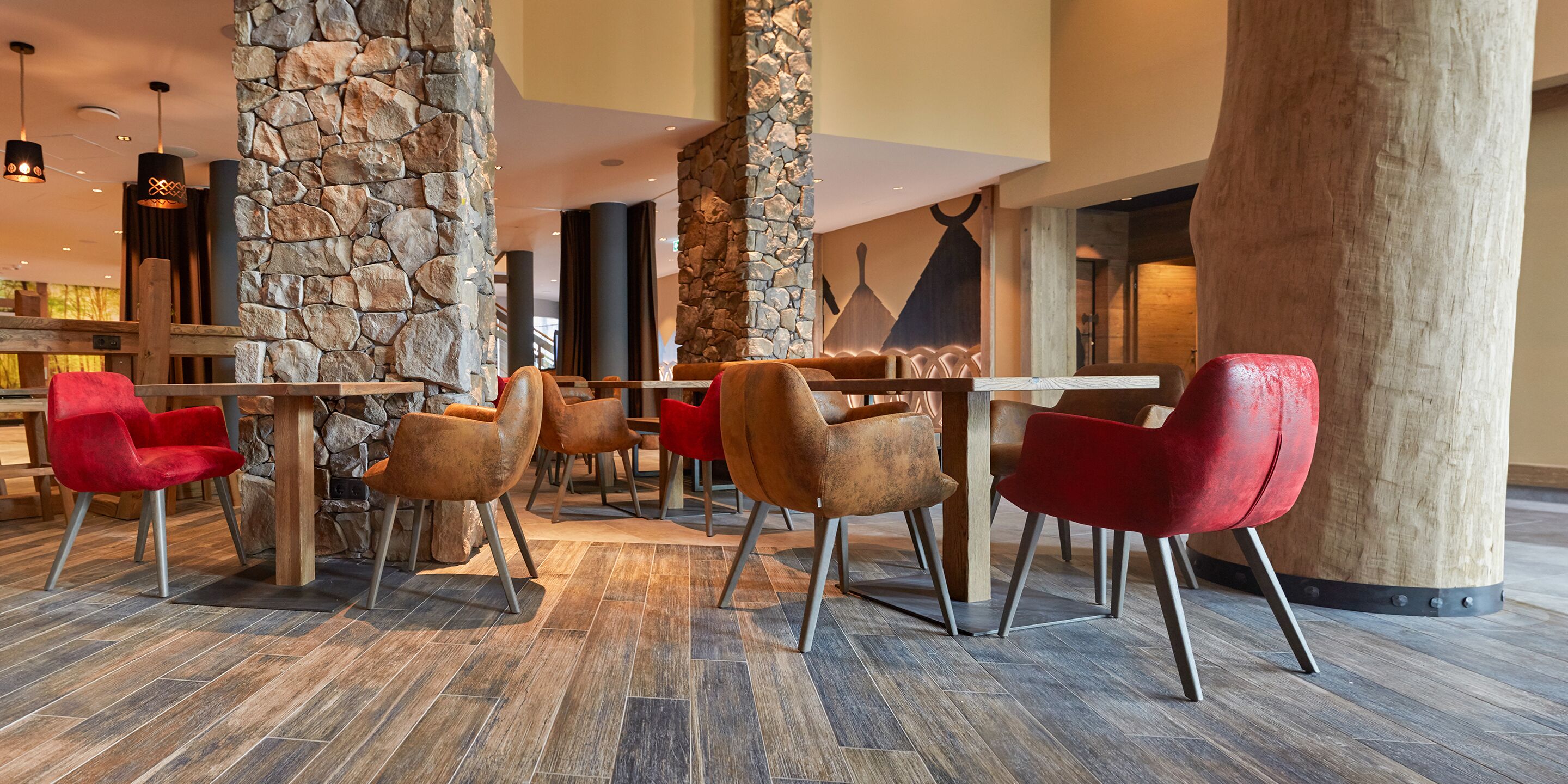 Un moderno vestíbulo con pilares de piedra, mesas de madera y sillones tapizados en tonos rojo y marrón.