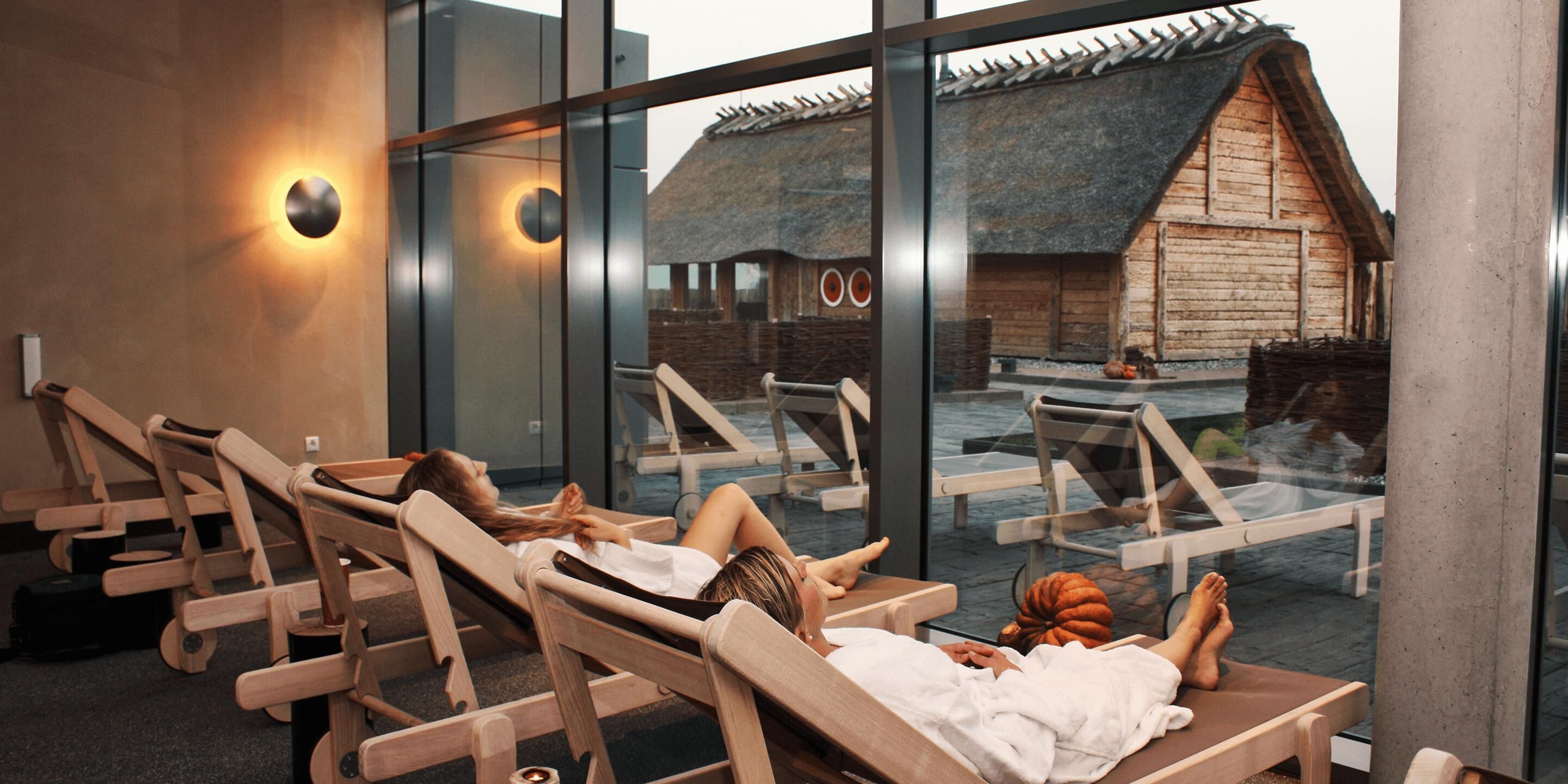 Un espace de détente avec des chaises longues en bois et un bâtiment à toit de chaume visible à travers des murs de verre.