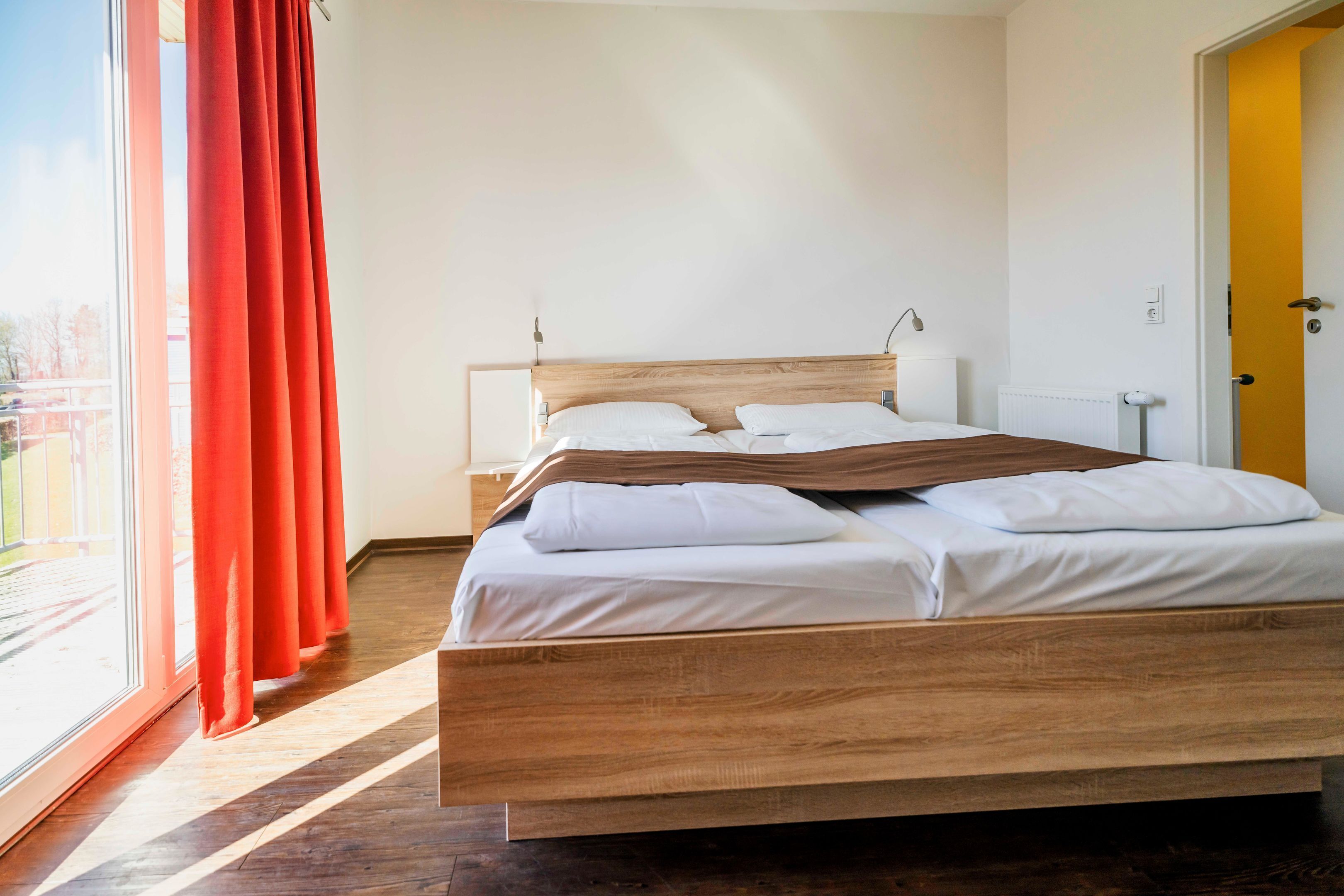 A bedroom with a wooden bed, white bedding, a nightstand, a lamp, and a view through a window with red curtains.