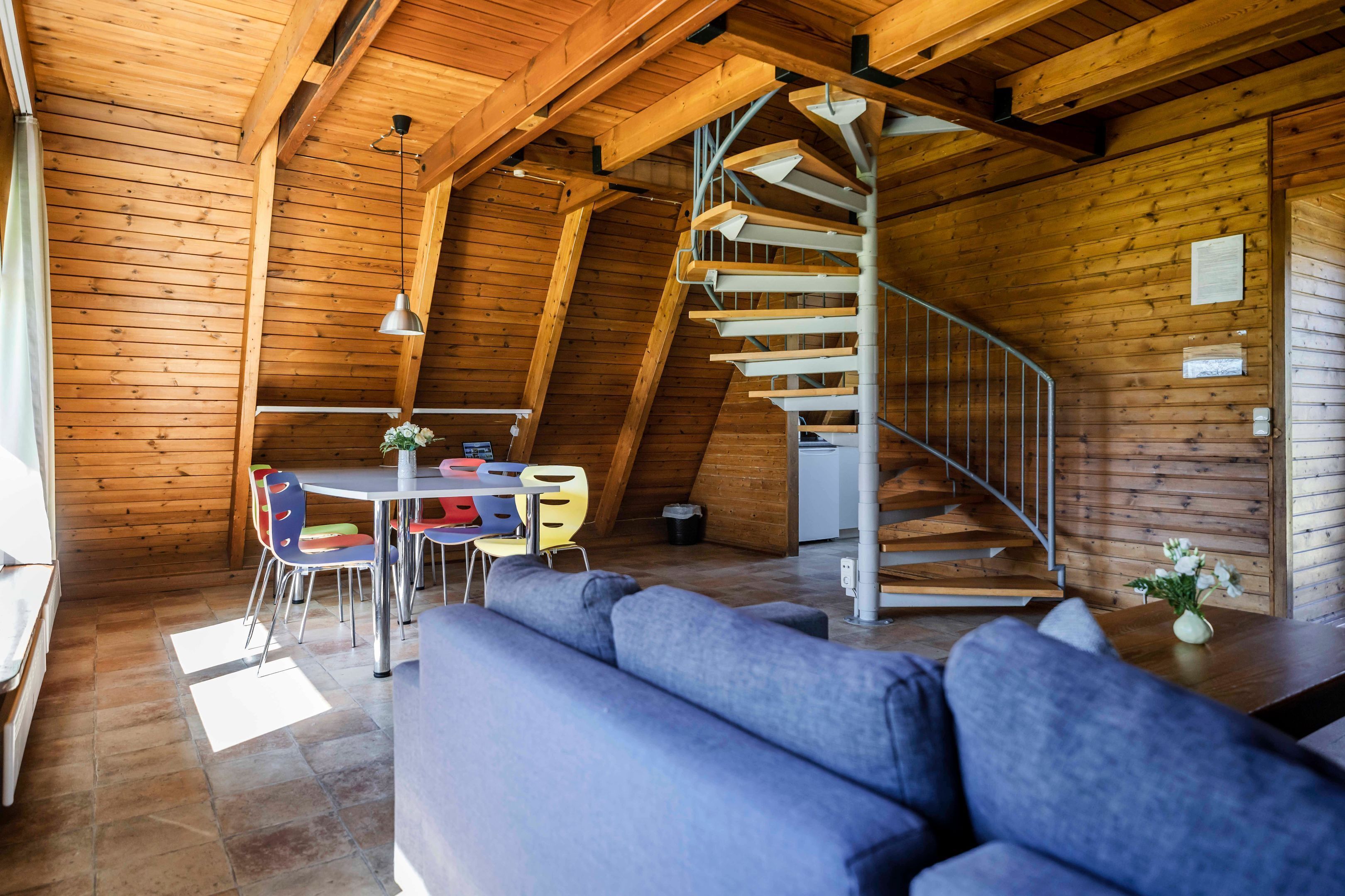 Ein Holzinnenraum mit einer Wendeltreppe, einem Essbereich mit bunten Stühlen und einem grauen Sofa.