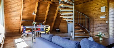 Een houten interieur met een wenteltrap, een eetgedeelte met kleurrijke stoelen en een grijze bank.